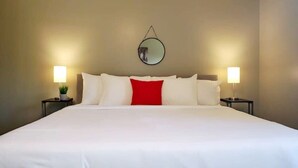 Una cama bien hecha con almohadas blancas y rojas, dos lámparas de noche y un espejo redondo en la pared.