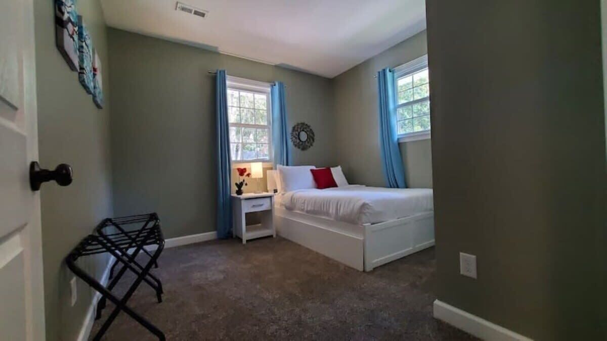 Quarto com cama, mesa de cabeceira, relógio na parede e janela com cortinas.
