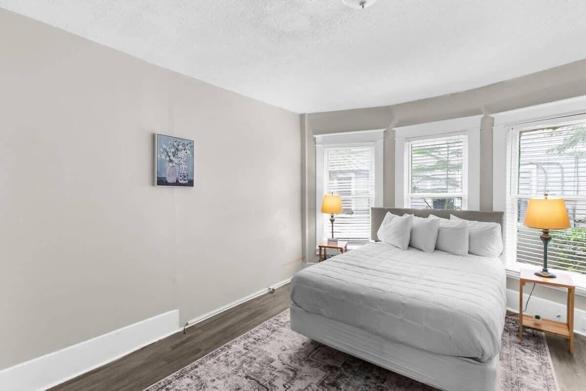 A bedroom with a large bed, two bedside tables with lamps, a window with blinds, and a framed picture on the wall.