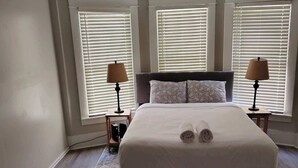 A neatly made bed with a rolled towel, two bedside lamps, and a patterned rug in a room with three windows and a wooden floor. - 29th Street Ensley Heights Hideaway 1 (Birmingham)