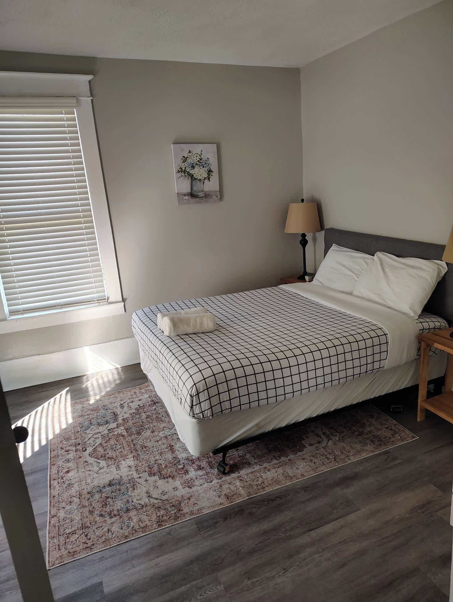 A bedroom with a bed, a nightstand, a lamp, a painting, and a window with blinds.