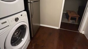 A laundry area with a front-loading washer and dryer, a refrigerator, and a wooden bench in the adjacent room.