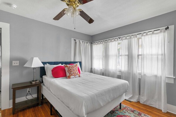 A bedroom with a bed, a nightstand, a lamp, a ceiling fan, and sheer curtains.