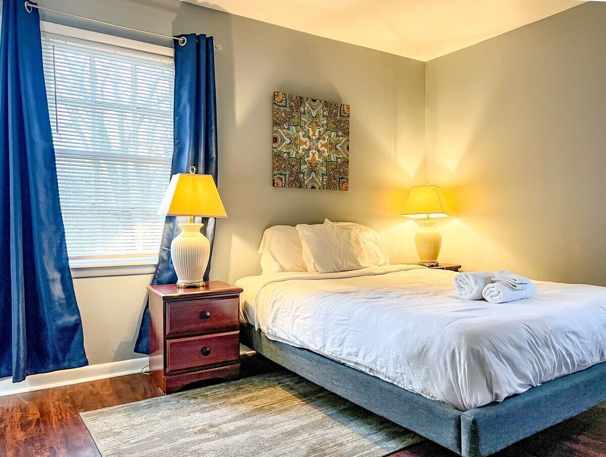 Een netjes ingerichte slaapkamer met een bed, twee bedlampen, een klein houten nachtkastje, een decoratief wandobject en een raam met blauwe gordijnen.