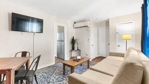 Uma sala de estar com sofá, mesa de centro de madeira, TV fixada na parede e área de cozinha visível por uma porta aberta.