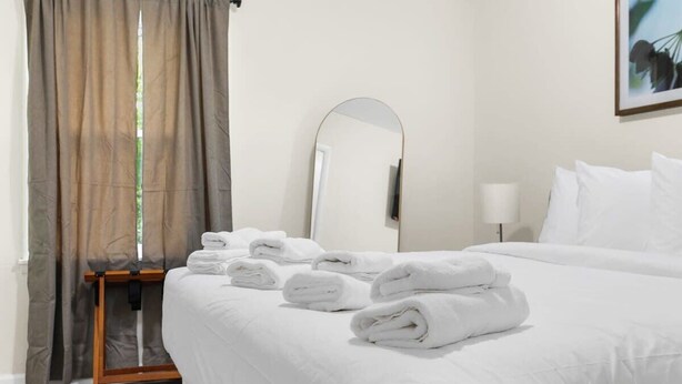 Cama bem arrumada com roupa de cama branca e toalhas dobradas, mesa de cabeceira, espelho e janela com cortinas.