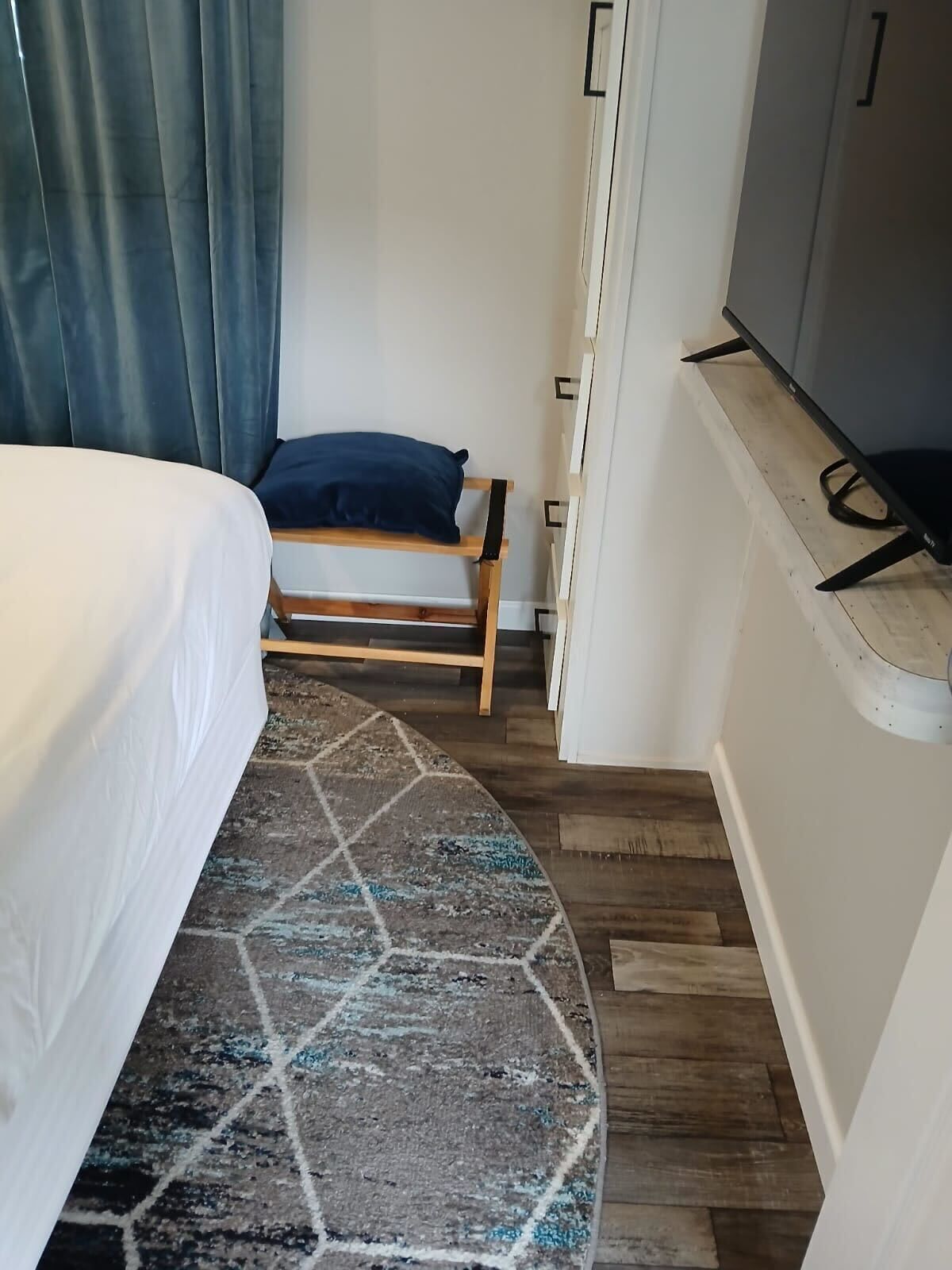 Una habitación pequeña y funcional con cama, banco de madera, televisor y alfombra estampada.