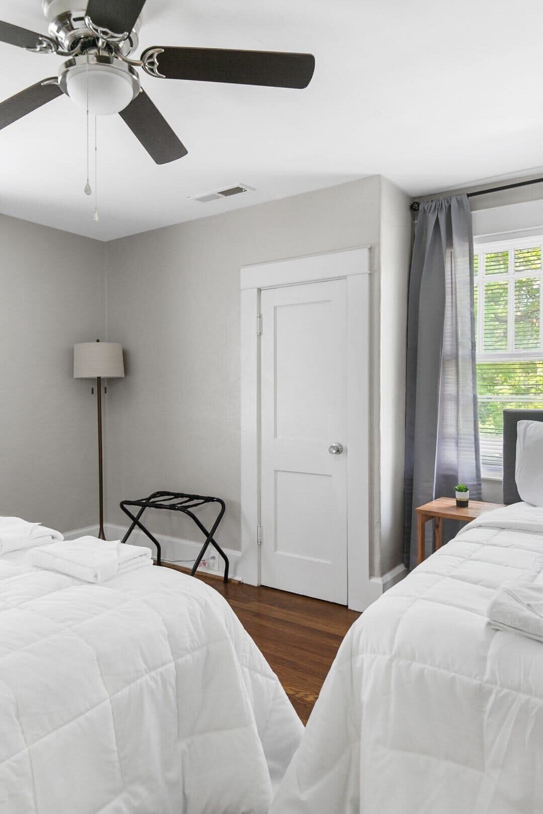 A hotel room with two beds, a ceiling fan, a door, a lamp, and a window with curtains.
