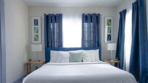 Een net opgemaakt bed met witte lakens, een blauw hoofdbord en twee nachtkastjes met lampen.