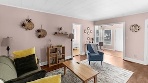 Een woonkamer met een bank, salontafel en een blauwe fauteuil. Er is een boekenkast met decoratieve objecten en een lamp. De kamer heeft een groot raam en een deur die naar buiten leidt.