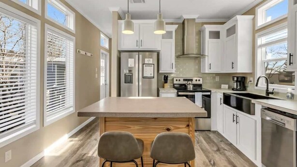 Une cuisine moderne dotée d’un îlot central, de meubles blancs, d’appareils électroménagers en acier inoxydable et de grandes fenêtres.