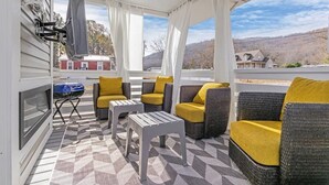 Un espace extérieur aménagé, abrité et doté de meubles en osier et de coussins, ainsi que d’un barbecue, offrant une vue sur une maison et des collines.