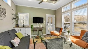 Un salon moderne avec un canapé, deux fauteuils, une table basse et un téléviseur. On y trouve des fenêtres équipées de stores, un ventilateur de plafond et, au fond, un coin cuisine.