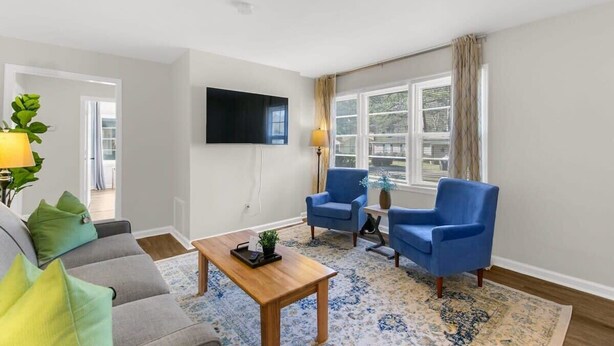 A living room with a sofa, two armchairs, a coffee table, a TV, and a large window with curtains.