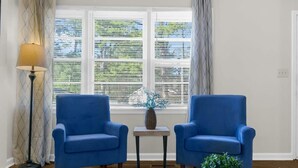 A room with two blue armchairs, a small table with a vase, a lamp, and a window with blinds.