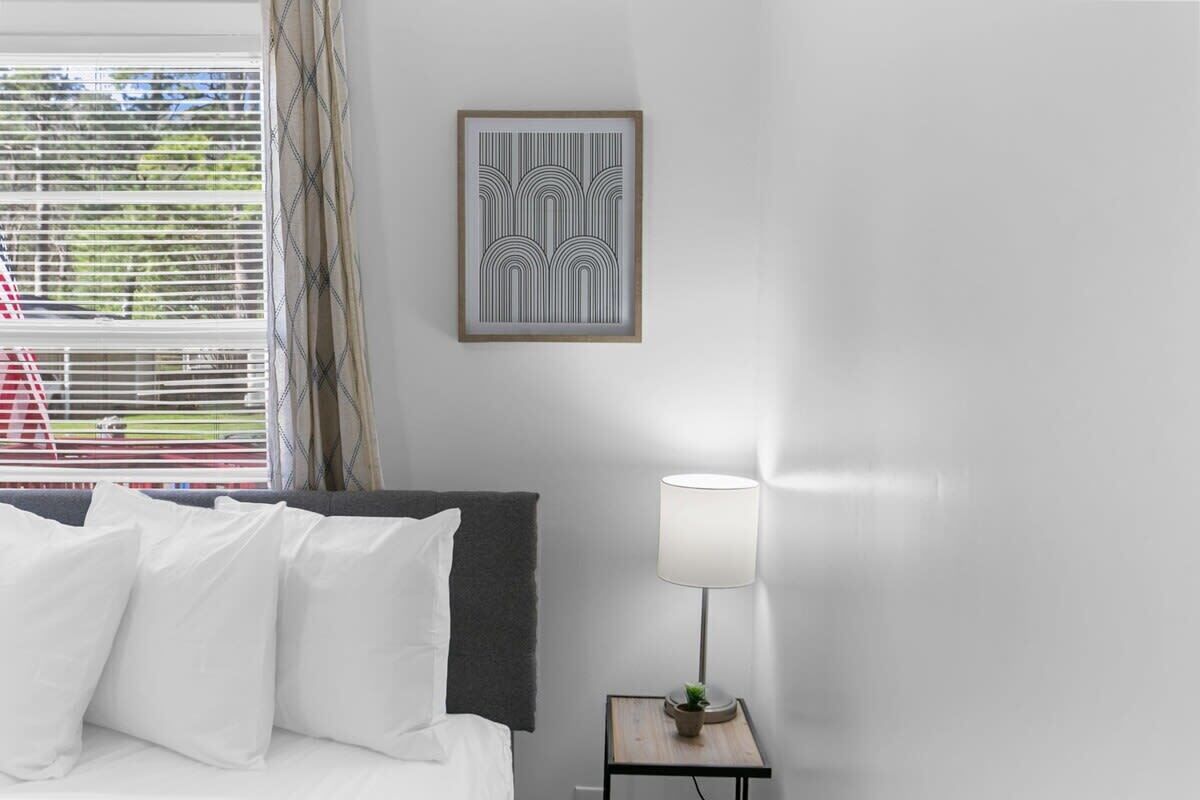 Habitación de hotel con cama, almohadas blancas, un cuadro en la pared, una lámpara de noche y una ventana con persianas.