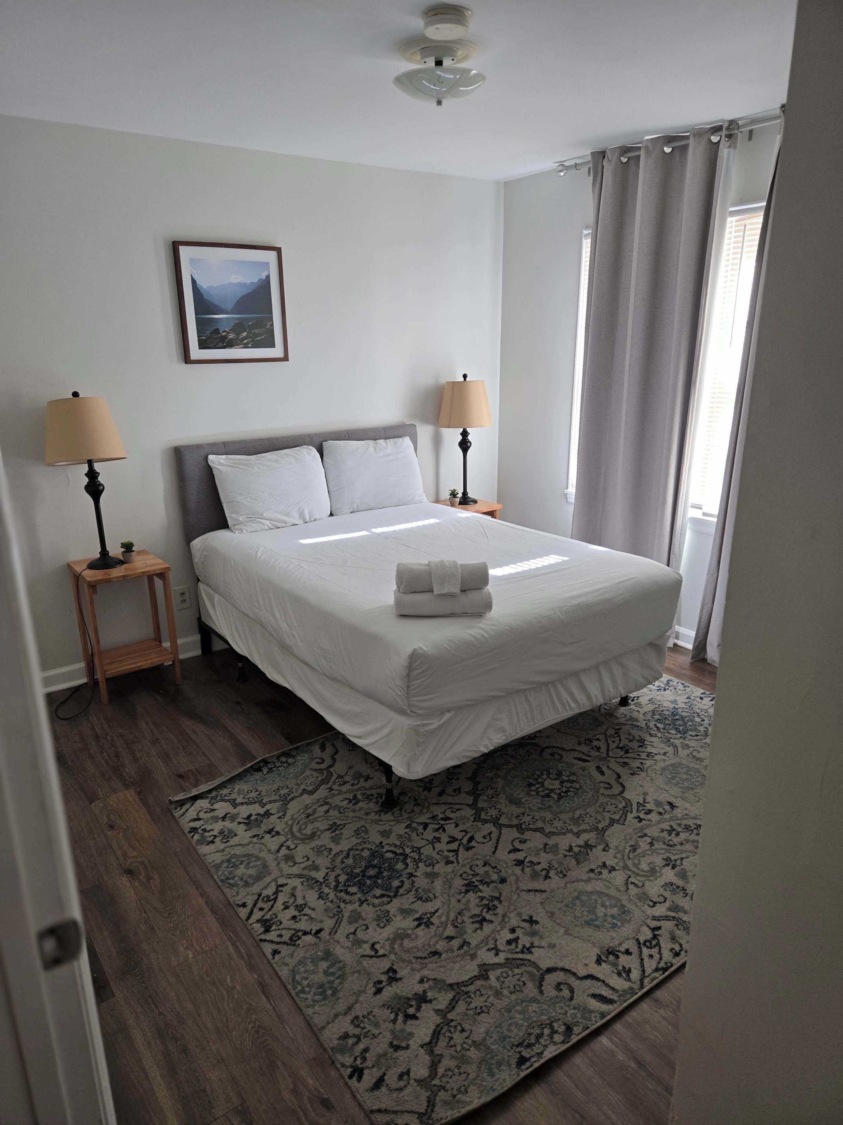 A neatly arranged bedroom with a bed, two bedside lamps, a framed picture on the wall, and a patterned rug.