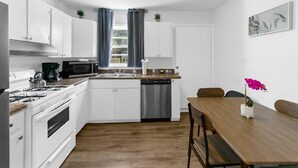 A compact kitchen with white cabinets, a microwave, a stove, and a dishwasher. There is a wooden dining table with chairs and a potted orchid.