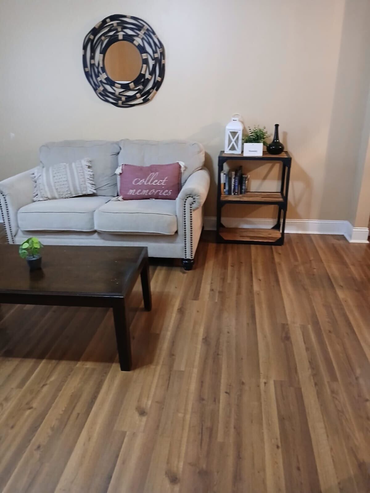A living room with a grey sofa, a black coffee table, a wooden floor, a decorative mirror, and a black bookshelf with books and a vase.