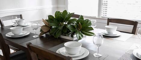 A dining table set with white plates, bowls, and wine glasses, surrounded by wooden chairs.