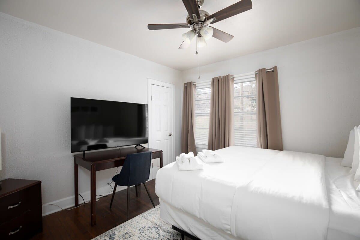 A hotel room with a bed, a desk with a TV, a ceiling fan, and a window with curtains.
