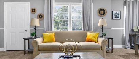 A living room with a beige sofa, two side tables with lamps, a coffee table, and a window with curtains.