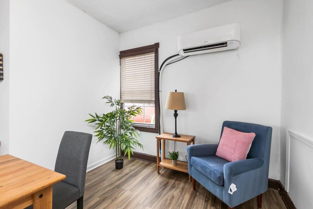 A room with a wooden table, a grey chair, a blue sofa, a small wooden side table with a lamp, a potted plant, and an air conditioning unit mounted on the wall.
