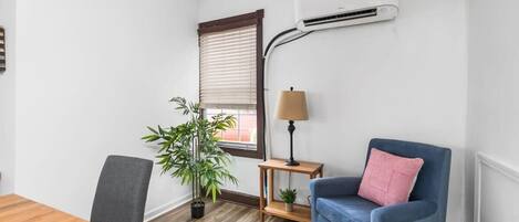 A room with a wooden table, a grey chair, a blue sofa, a small wooden side table with a lamp, a potted plant, and an air conditioning unit mounted on the wall.