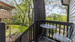 Une terrasse en bois avec une table et une chaise en métal, entourée d’arbres et d’une zone herbeuse.