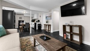 A modern living room with a sofa, coffee table, and a TV mounted on the wall. There is a kitchen area in the background with a dining table and chairs. - Dugan Ave Unit B (Birmingham)