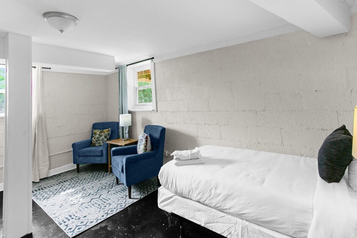 Quarto de hotel moderno com uma cama grande, uma poltrona azul, uma mesinha de cabeceira com abajur e vista para o exterior pela janela.