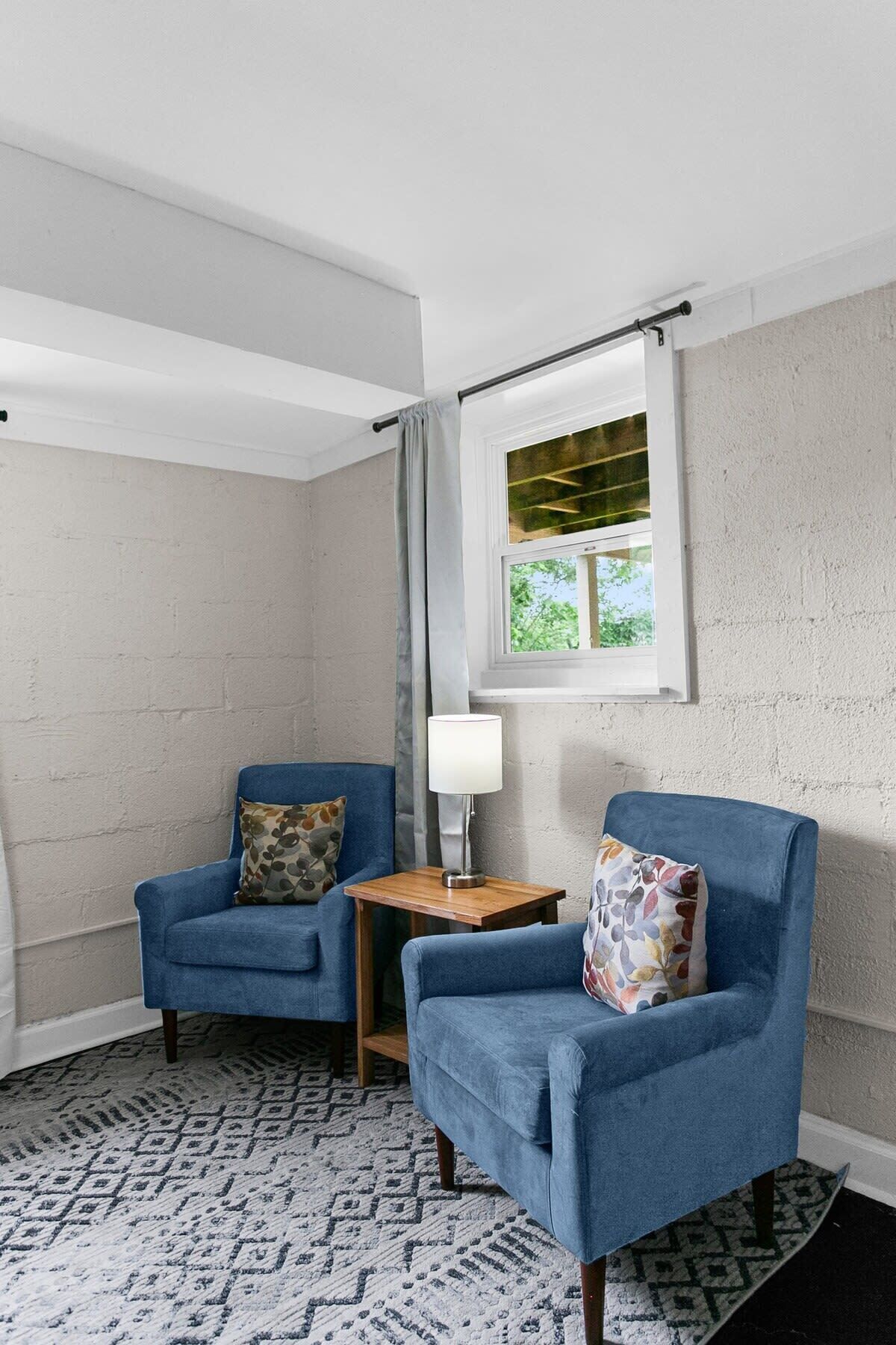 A room with two blue armchairs, a small wooden table with a lamp, and a window with curtains.