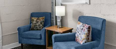 A room with two blue armchairs, a small wooden table with a lamp, and a window with curtains.