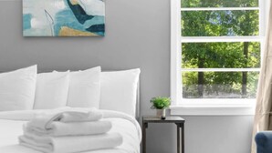 A neatly made bed with white linens, a small bedside table with a plant, and a window with a view of trees. - Broadmoor Bungalow - Unit A (Birmingham)