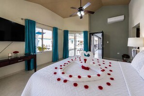 A hotel room with a bed adorned with rose petals and a heart-shaped swan decoration, a ceiling fan, a flat-screen TV, and a view of the outdoor area.