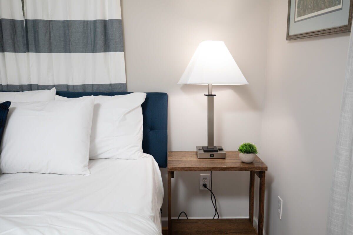 A neatly made bed with white linens and a blue headboard, a bedside table with a lamp, and a small potted plant.
