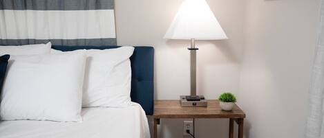 A neatly made bed with white linens and a blue headboard, a bedside table with a lamp, and a small potted plant.