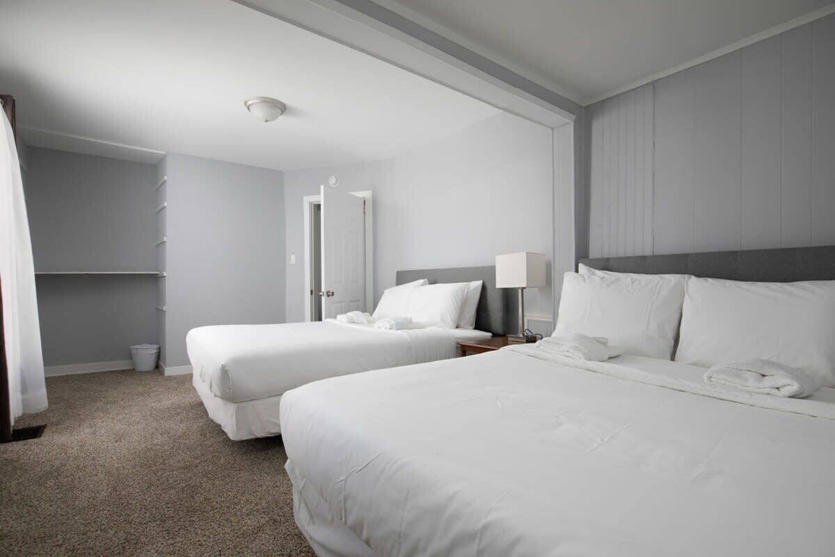 A hotel room with two beds, a bedside table with a lamp, a wardrobe, and a carpeted floor.