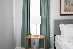 A neatly arranged bedroom with a bed, a bedside table with a lamp, and a window with green curtains. - 4th St Little Blue in Bham (Birmingham)