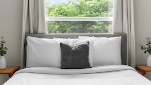 A bed with white bedding and a grey pillow, a window with curtains, and a view of trees. - Dugan Ave Both (Birmingham)