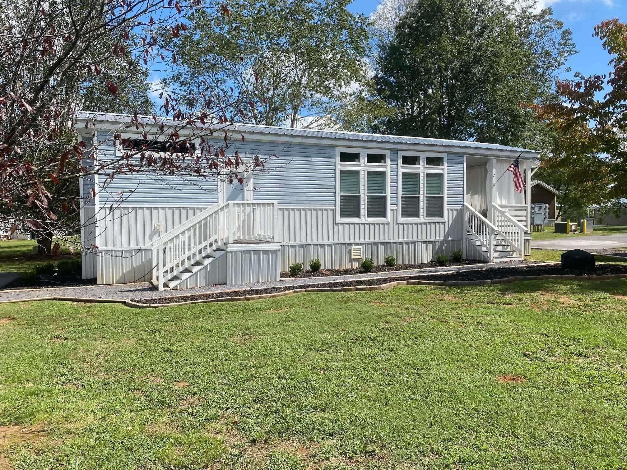 A single-story, light blue mobile home with a white exterior, a front porch with a staircase, and an American flag on the right side.