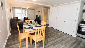 Un coin repas avec une table et des chaises en bois, un téléviseur en arrière-plan et un espace cuisine équipé d’une cuisinière.
