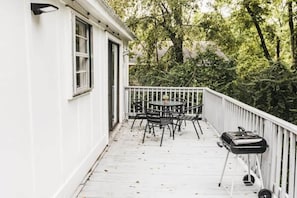 Une terrasse en bois peinte en blanc, avec une table et des chaises, entourée d’une balustrade blanche, et donnant sur une zone boisée.