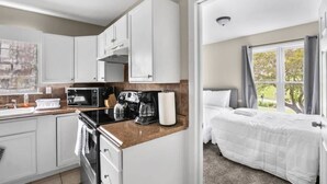 Eine kleine Küche mit weißen Schränken, einer Mikrowelle, einem Wasserkocher und einer Kaffeemaschine. Angrenzend befindet sich ein Schlafzimmer mit Bett, Fenster und Vorhängen.