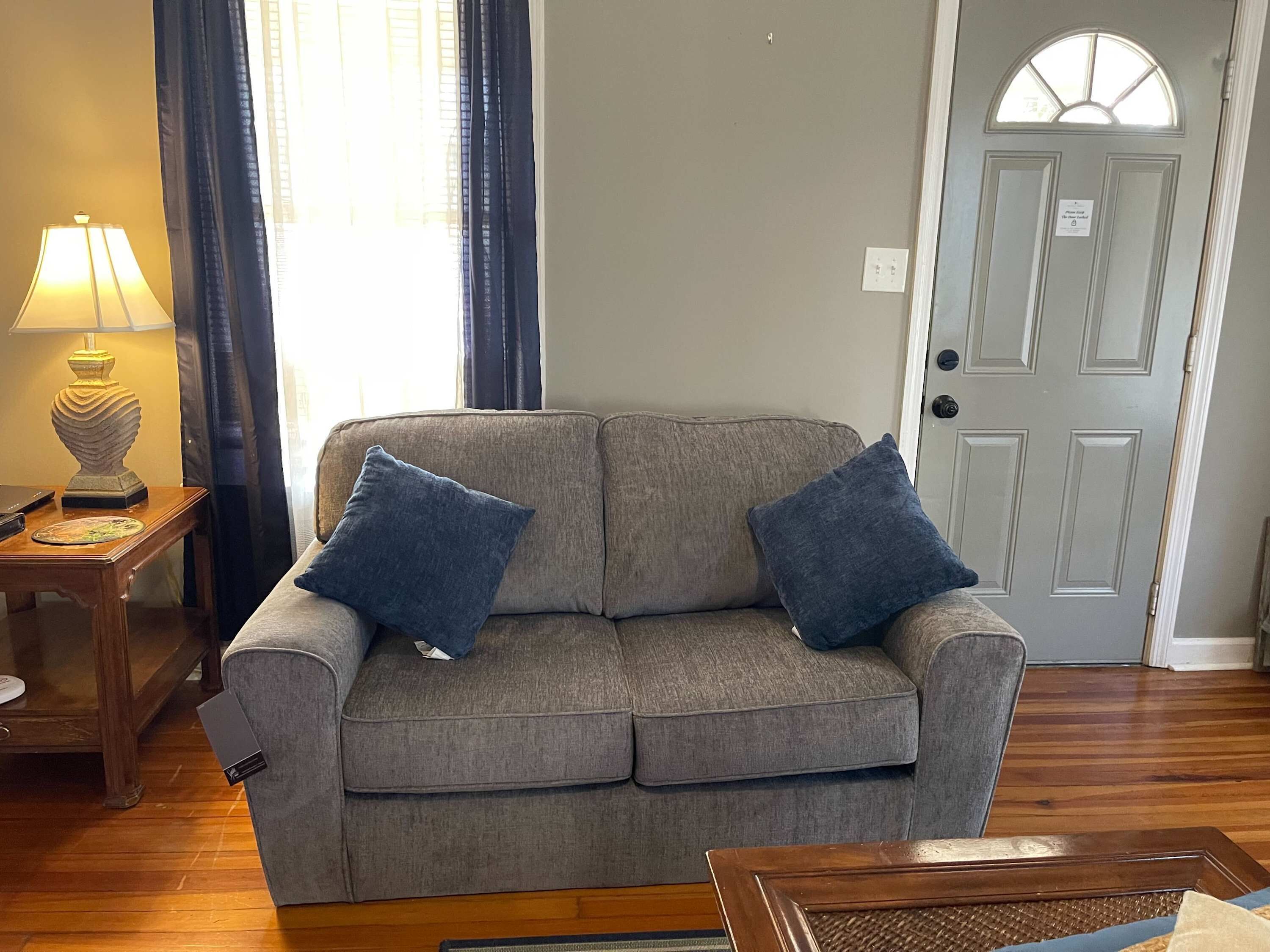 A gray sofa with two blue cushions, a wooden side table with a lamp, and a door with a sign.