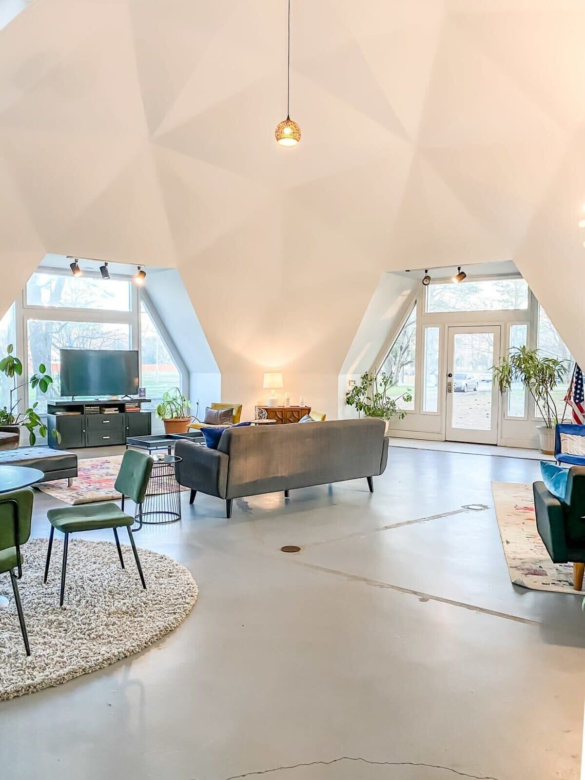A spacious loft-style living area with a high ceiling, a central seating area with a sofa and chairs, a television, and a dining table with chairs.