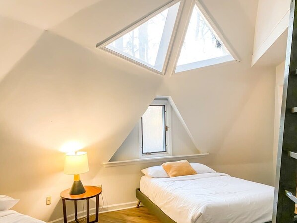 A bedroom with a sloped ceiling, skylight windows, a bed with white bedding, and a bedside table with a lamp.