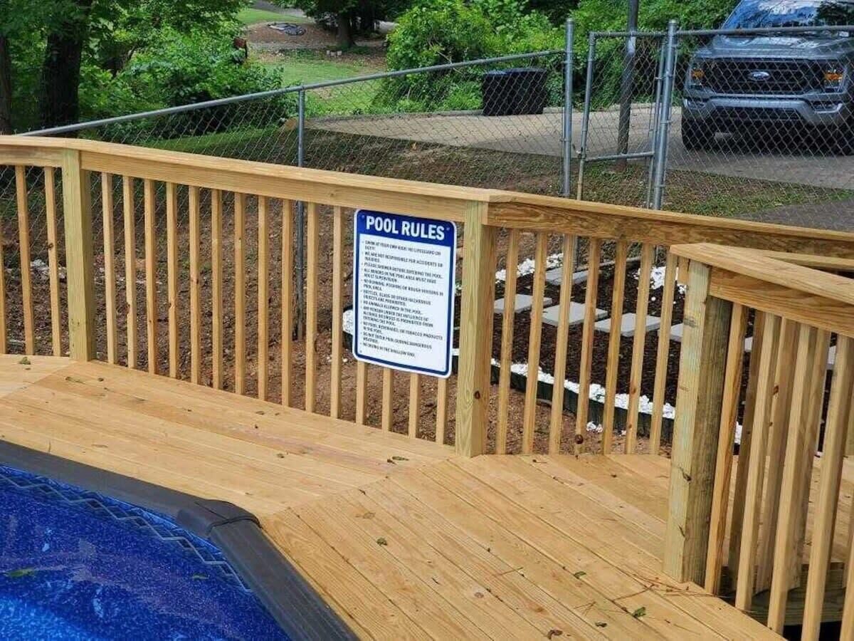Una terraza de madera con piscina, un letrero con las normas de la piscina y una valla al fondo.