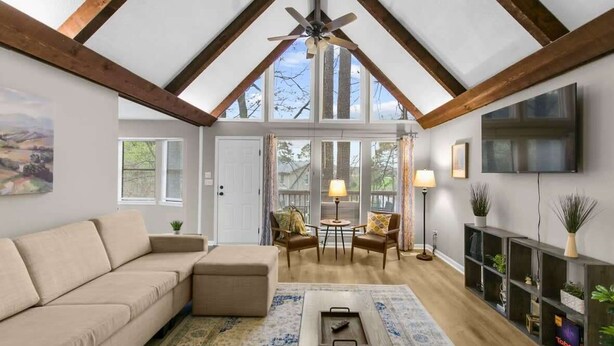 A spacious living room with a high ceiling, wooden beams, a large sofa, a coffee table, two chairs, a TV, and a bookshelf. - Valley Lake Gem (Birmingham)