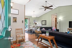Ein Wohnzimmer mit einer Couch, Sesseln, einem Couchtisch und einem Fernseher. Es gibt einen Deckenventilator, eine Chandelier und eine Tür mit einem Spiegel.